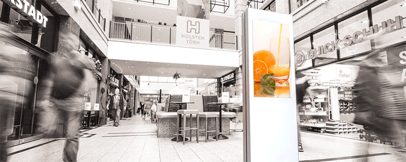 Ein Digital Signage Bildschirm in einem Einkaufszentrum. Es läuft ein Video, in welchem ein Glas Orangensaft eingeschenkt wird.